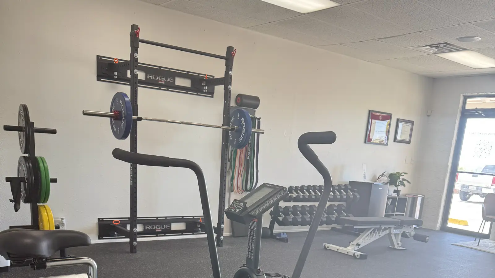 The training floor at Solas Physical Therapy, El Paso: Rogue rack with a loaded barbell and bumper plates, dumbbells and an assault bike
