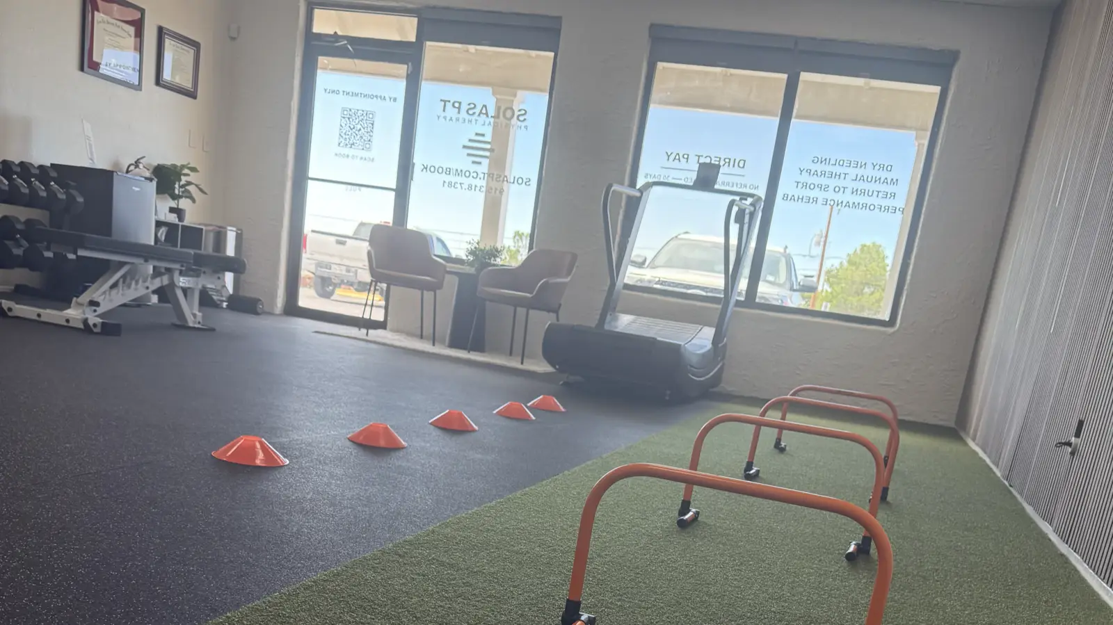 Return-to-sport testing setup on the turf lane at Solas Physical Therapy, El Paso: agility hurdles and cones laid out in the open one-on-one treatment room, with the curved treadmill and dumbbells behind